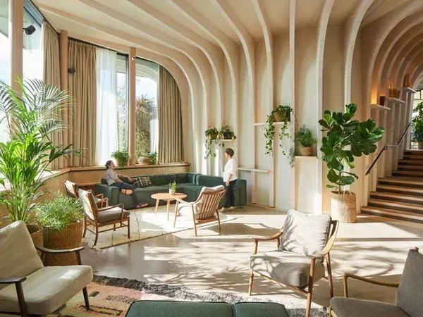 A modern lounge interior featuring curved exposed timber beams, natural light from tall windows, and abundant indoor greenery. People are seated and standing, emphasizing a relaxed, biophilic atmosphere